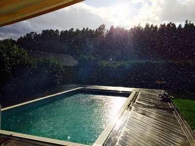 A swimming pool is surrounded by a wooden deck, with droplets of water visible in the air. The sun is shining brightly through a partly cloudy sky, casting reflections on the water. In the background, a dense line of trees provides a natural border.