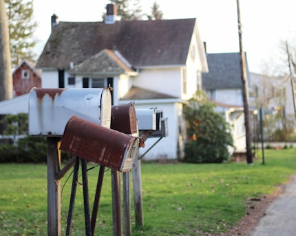 A row of old, rusted mailboxes on wooden posts stands in front of a house with a sloped roof. The house is set in a suburban neighborhood with green grass and a gravel path nearby. The scene is calm and somewhat nostalgic.