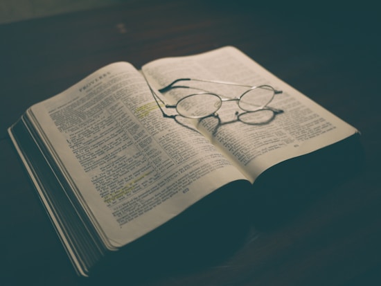 An open book lays on a wooden table with reading glasses resting on one of its pages. The lighting is dim, creating a warm and vintage atmosphere. The text in the book is highlighted in places with a yellow marker, suggesting important sections.