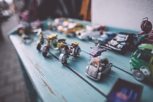 A collection of colorful, miniature toy vehicles is arranged on a weathered, teal-painted wooden table. The toys include various styles such as scooters, motorcycles, and cars, all depicted in a vintage style with intricate details. The focus is on the foreground with the background toys softly blurred, creating a depth-of-field effect. The setting appears casual and outdoors.