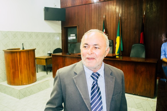 A man with a white beard and short hair is wearing a suit and striped tie. He is standing in what appears to be a courtroom or formal meeting room with wooden walls and furniture. There are flags in the background and a microphone on the podium.