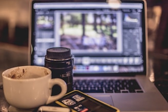 A coffee cup is placed next to a camera lens and a laptop displaying a digital photo editing software. The background is slightly blurred, giving focus to the items on the table, including an SD card holder and a phone.