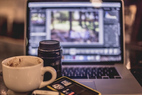 A coffee cup is placed next to a camera lens and a laptop displaying a digital photo editing software. The background is slightly blurred, giving focus to the items on the table, including an SD card holder and a phone.
