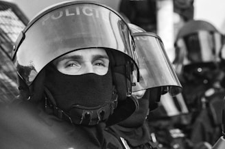 A black-and-white photograph of individuals wearing protective gear and helmets, capturing the serious expression of a person in the foreground. The visor of the helmet reflects light, and the word 'POLICE' is visible. Other individuals similarly outfitted are present in the background.