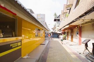 A narrow street with closed shops on either side, where the left side features a bright yellow Western Union building. There are a few people walking along the street under a clear sky. Some of the buildings have signs in different languages.