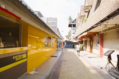 A narrow street with closed shops on either side, where the left side features a bright yellow Western Union building. There are a few people walking along the street under a clear sky. Some of the buildings have signs in different languages.