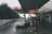 A sleek white sports car is parked at a gas station, under a large canopy with bold orange and white signage. The station is situated in a wooded area, with trees in the background and a misty sky, creating a calm atmosphere. A wet road is visible beside the gas station.