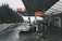 A sleek white sports car is parked at a gas station, under a large canopy with bold orange and white signage. The station is situated in a wooded area, with trees in the background and a misty sky, creating a calm atmosphere. A wet road is visible beside the gas station.