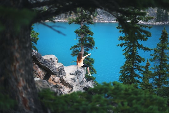 girl sitting on cliff near trees pointing towards body of water in Moraine Lake Canada