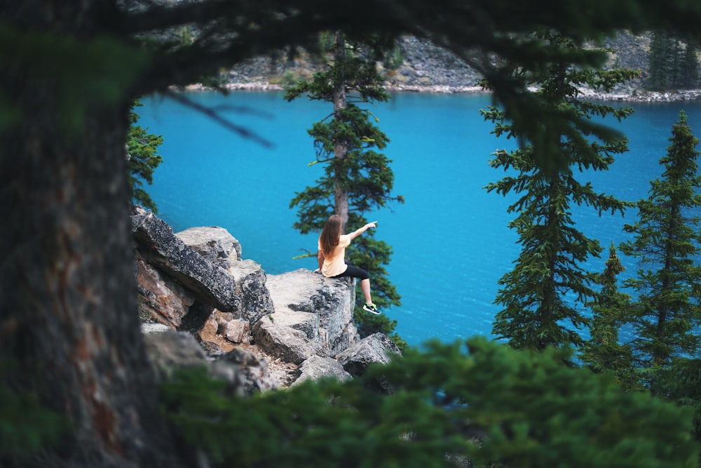 girl sitting on cliff near trees pointing towards body of water