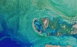 Aerial view of a coastal area with swirling green and blue hues, representing the ocean or sea. The central part features small land masses or islands surrounded by shallow waters, creating intricate patterns. The coastline is visible at the bottom, with sandy and rocky textures.