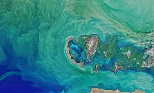 Aerial view of a coastal area with swirling green and blue hues, representing the ocean or sea. The central part features small land masses or islands surrounded by shallow waters, creating intricate patterns. The coastline is visible at the bottom, with sandy and rocky textures.