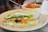 A fresh vegetable sandwich is placed on a plate with a toothpick securing the layers. The sandwich contains sliced cucumbers, shredded carrots, olives, tomatoes, lettuce, and cream cheese spread, all between two thick slices of bread. Behind it, a white bowl filled with tomato soup is partially visible, garnished with herbs and featuring a spoon inside.