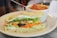 A fresh vegetable sandwich is placed on a plate with a toothpick securing the layers. The sandwich contains sliced cucumbers, shredded carrots, olives, tomatoes, lettuce, and cream cheese spread, all between two thick slices of bread. Behind it, a white bowl filled with tomato soup is partially visible, garnished with herbs and featuring a spoon inside.
