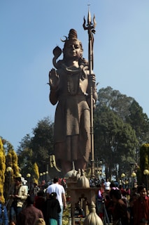 A large, intricate statue of a deity holding a trident stands tall surrounded by trees and a crowd of people. The sky is clear, providing a vivid blue backdrop. The statue features detailed craftsmanship, including a snake around its neck and a serene expression.