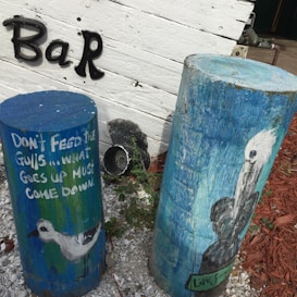 Two painted wooden posts with illustrations of birds. One post features a painted seagull and the phrase 'DON'T FEED THE GULLS...WHAT GOES UP MUST COME DOWN.' The backdrop includes a white wooden wall with the word 'Bar' spelled out in metal letters. The ground is covered in gravel and mulch, and there's a small bush nearby.