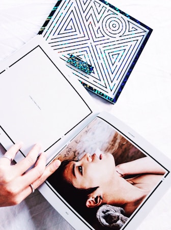 A hand holding open a photo book on a white surface. The open page displays a close-up image of a person looking upwards with a dreamy expression. Next to the book, a colorful item with geometric patterns is partially visible.