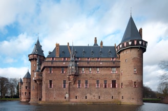 A large medieval-style castle made of brick with several towers, each topped with conical dark gray roofs. The castle is surrounded by a moat, reflecting part of the structure. Windows are adorned with decorative red and white shutters. The sky is partly cloudy, creating a mix of sunlight and shadows on the castle walls.