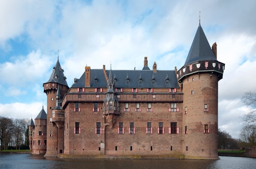 A large medieval-style castle made of brick with several towers, each topped with conical dark gray roofs. The castle is surrounded by a moat, reflecting part of the structure. Windows are adorned with decorative red and white shutters. The sky is partly cloudy, creating a mix of sunlight and shadows on the castle walls.