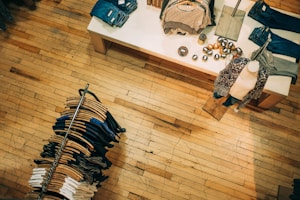 A retail clothing store with a wooden floor. A display table shows neatly folded clothes and decorative items, while a mannequin is dressed in patterned clothing. A circular clothing rack holds various-colored garments on hangers.