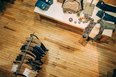 A retail clothing store with a wooden floor. A display table shows neatly folded clothes and decorative items, while a mannequin is dressed in patterned clothing. A circular clothing rack holds various-colored garments on hangers.