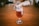 A glass filled with amber-colored beer sits on a wooden table, partially frothy on top. The background is blurred, showing a warm, indoor setting with additional glasses out of focus.