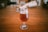 A glass filled with amber-colored beer sits on a wooden table, partially frothy on top. The background is blurred, showing a warm, indoor setting with additional glasses out of focus.