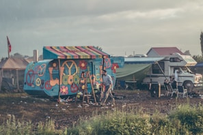 A vibrantly painted camper van displaying peace symbols and flower motifs is situated in a relatively open field. Beside it is a more traditional motorhome with a green tarp extending from its side. Various camping chairs are arranged nearby, and a person is walking through the area. The atmosphere appears somewhat rustic with scattered litter on the ground and a flag displayed on a tent in the background.