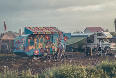 A vibrantly painted camper van displaying peace symbols and flower motifs is situated in a relatively open field. Beside it is a more traditional motorhome with a green tarp extending from its side. Various camping chairs are arranged nearby, and a person is walking through the area. The atmosphere appears somewhat rustic with scattered litter on the ground and a flag displayed on a tent in the background.