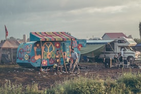 A vibrantly painted camper van displaying peace symbols and flower motifs is situated in a relatively open field. Beside it is a more traditional motorhome with a green tarp extending from its side. Various camping chairs are arranged nearby, and a person is walking through the area. The atmosphere appears somewhat rustic with scattered litter on the ground and a flag displayed on a tent in the background.