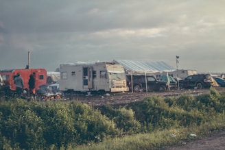 A campsite with several RVs and tents alongside parked vehicles. Two figures are standing by a small campfire, and there's an overcast sky above. The scene is set on a grassy area with visible greenery in the foreground.
