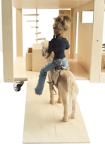 A doll is seated on a wooden horse, facing a minimalistic wooden structure with geometric shapes. The doll is wearing a navy jacket and blue jeans, and has short brown hair.