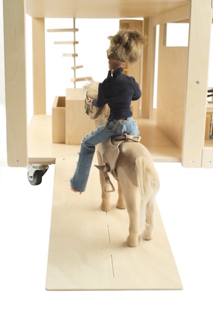 A doll is seated on a wooden horse, facing a minimalistic wooden structure with geometric shapes. The doll is wearing a navy jacket and blue jeans, and has short brown hair.