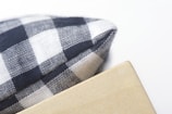 A close-up of the corner of a checkered fabric pillow next to a wooden surface. The fabric features a black and white checkered pattern, and the wooden surface is light in color, with visible grain.