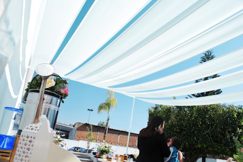 Several white fabric canopies are stretched overhead in an outdoor setting. A table is set up with various items, including a metal watering can filled with flowers. In the background, trees, a building, and a clear blue sky are visible. Two people, one holding a child, are present and appear to be engaged in conversation.