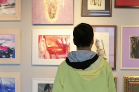 A person wearing a light jacket stands in front of a gallery wall filled with various framed artworks. The paintings and drawings vary in style, with some incorporating vibrant colors and others featuring more subdued tones. The artworks are displayed in an organized manner, with different sizes and types of frames.