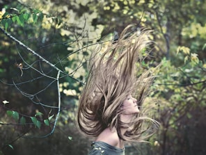 A person is seen with long hair dramatically flowing upwards, surrounded by lush, green foliage. The background consists of blurred green leaves with light filtering through, creating a natural and organic feel.