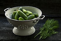 A white colander filled with fresh cucumbers sits on a dark wooden surface. Next to the colander is a bunch of fresh dill, adding a vibrant green contrast to the scene. The lighting casts a natural, rustic ambiance.