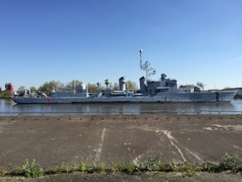 A large, grey military ship with various antenna and equipment on its deck, docked by a calm waterway. The ship is marked with the designation 'D627' on its side. In the background, there are trees and a small red building under a clear blue sky. The foreground features a concrete surface with some green vegetation along the edge.
