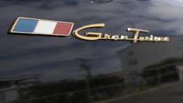 A close-up view of a logo with three vertical stripes in blue, white, and red next to the gold script 'Gran Turismo' on a dark surface.