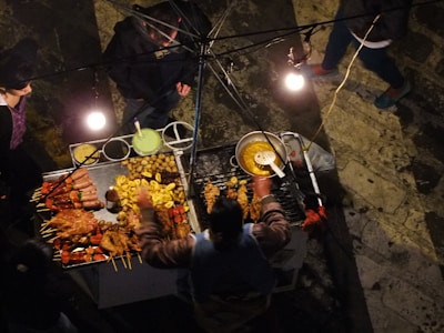 A top-down view of a street food vendor's stall, showcasing a variety of grilled meats on skewers alongside potatoes and other fried foods. The stall is illuminated by several light bulbs, creating a warm glow. People are gathered around, likely customers or the vendor.