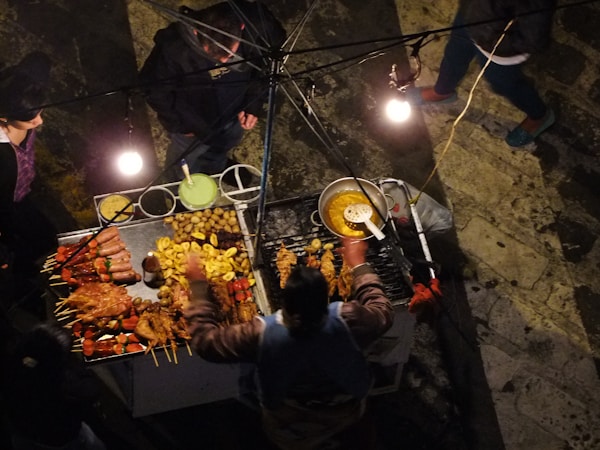 A top-down view of a street food vendor's stall, showcasing a variety of grilled meats on skewers alongside potatoes and other fried foods. The stall is illuminated by several light bulbs, creating a warm glow. People are gathered around, likely customers or the vendor.