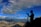 A silhouette of a person with a camera on a tripod stands against a vibrant blue sky filled with clouds and a vivid rainbow. The landscape below features expansive mountain ranges under soft sunlight.