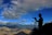 A silhouette of a person with a camera on a tripod stands against a vibrant blue sky filled with clouds and a vivid rainbow. The landscape below features expansive mountain ranges under soft sunlight.