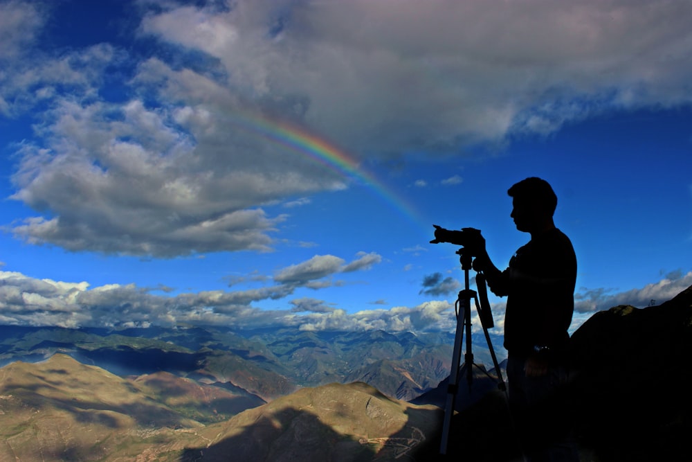 silhouette photo of holding camera with stand during daytime with rainbow