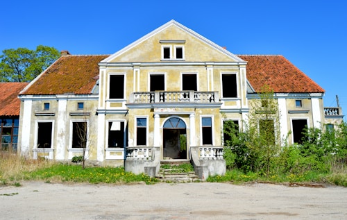 An abandoned, old two-story house with a dilapidated facade, featuring missing windows and overgrown plants. The roof has a reddish-brown hue, and the walls are a faded yellow with signs of deterioration. The foreground shows an unkempt yard with scattered grass and dirt patches.