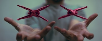 Two red origami figures hover in mid-air above a pair of outstretched hands. The background is softly blurred, focusing attention on the detailed paper creations.