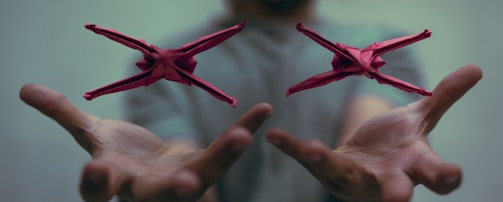 Two red origami figures hover in mid-air above a pair of outstretched hands. The background is softly blurred, focusing attention on the detailed paper creations.