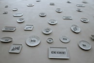 A collection of white ceramic plates and rectangular dishes displayed on a wall. Each piece has a German word or phrase printed in bold, black font. The words appear in various sizes and orientations, adding an abstract and artistic element to the display.