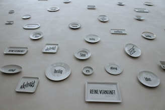 A collection of white ceramic plates and rectangular dishes displayed on a wall. Each piece has a German word or phrase printed in bold, black font. The words appear in various sizes and orientations, adding an abstract and artistic element to the display.